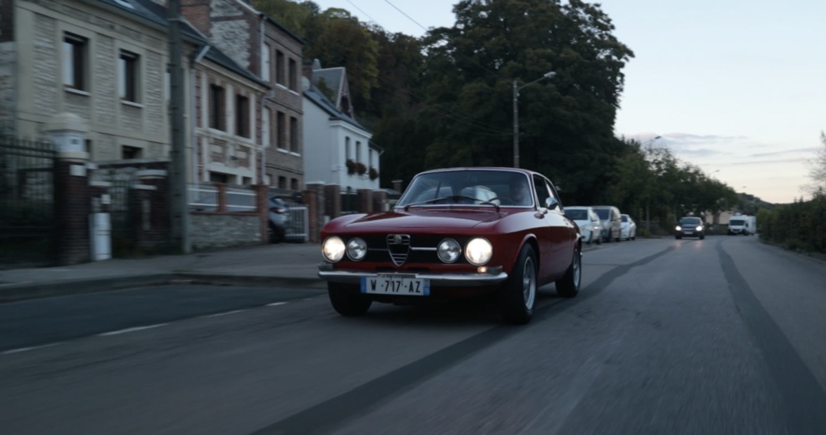 Alfa Romeo 1750 GTV rouge filmée de face sur une route, phares allumés, dans une atmosphère cinématographique douce et légèrement ancienne.